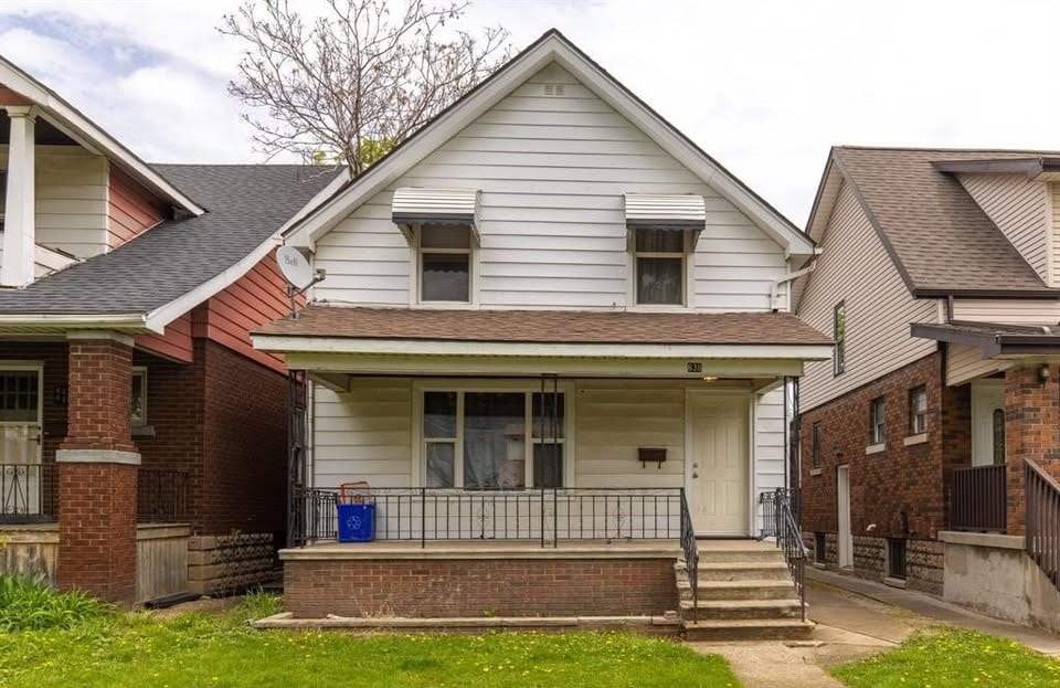 White clapboard two-storey house with a covered front porch, metal railings and awnings over the upper windows, between two brick neighbours.