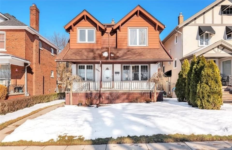 Two-storey house with rust-red gabled dormers, a white railed front porch and a snow-covered front lawn, on a street of similar detached homes in Windsor.