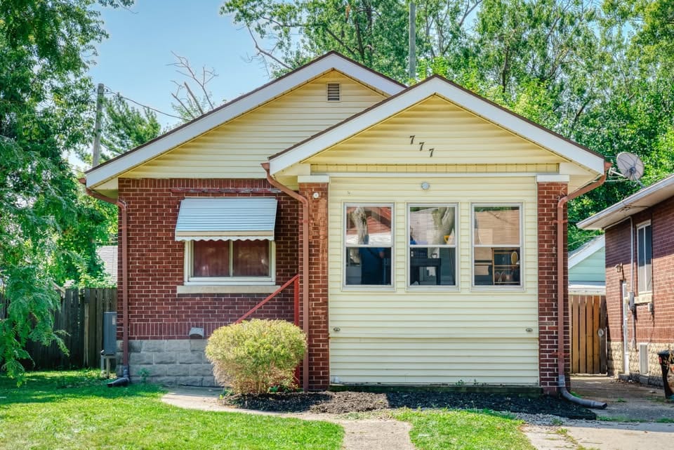 Single-storey bungalow with pale yellow siding and red brick, a wide front picture window and a shrub beside the front path, photographed in summer.